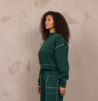 A woman stands in profile wearing a dark green knit matching set consisting of a long-sleeve cropped sweatshirt and wide-leg pants with contrasting light beige stitching. She is wearing beige sneakers and standing against a textured, warm-toned concrete wall.