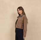 A woman stands in profile wearing a cropped brown jacket with a dark collar, black wide-leg pants, and black shoes against a warm beige textured wall.