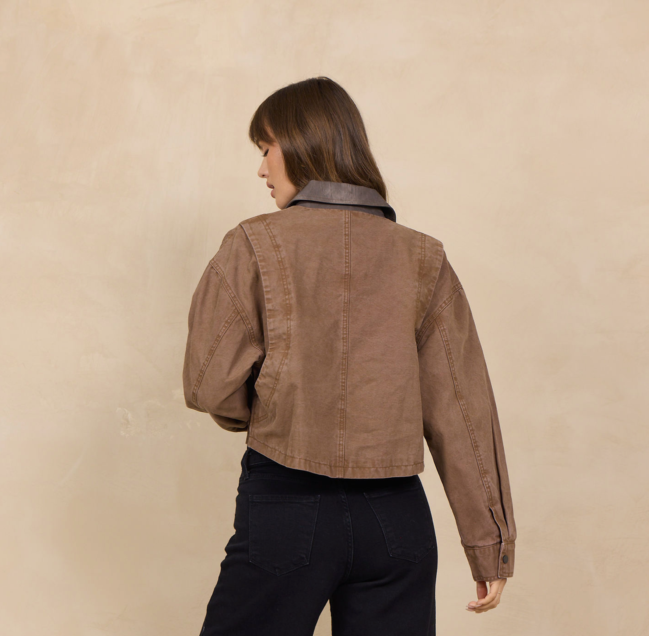 A woman is shown from the back wearing a cropped brown jacket with a dark gray collar and black wide-leg pants, standing against a textured beige wall.