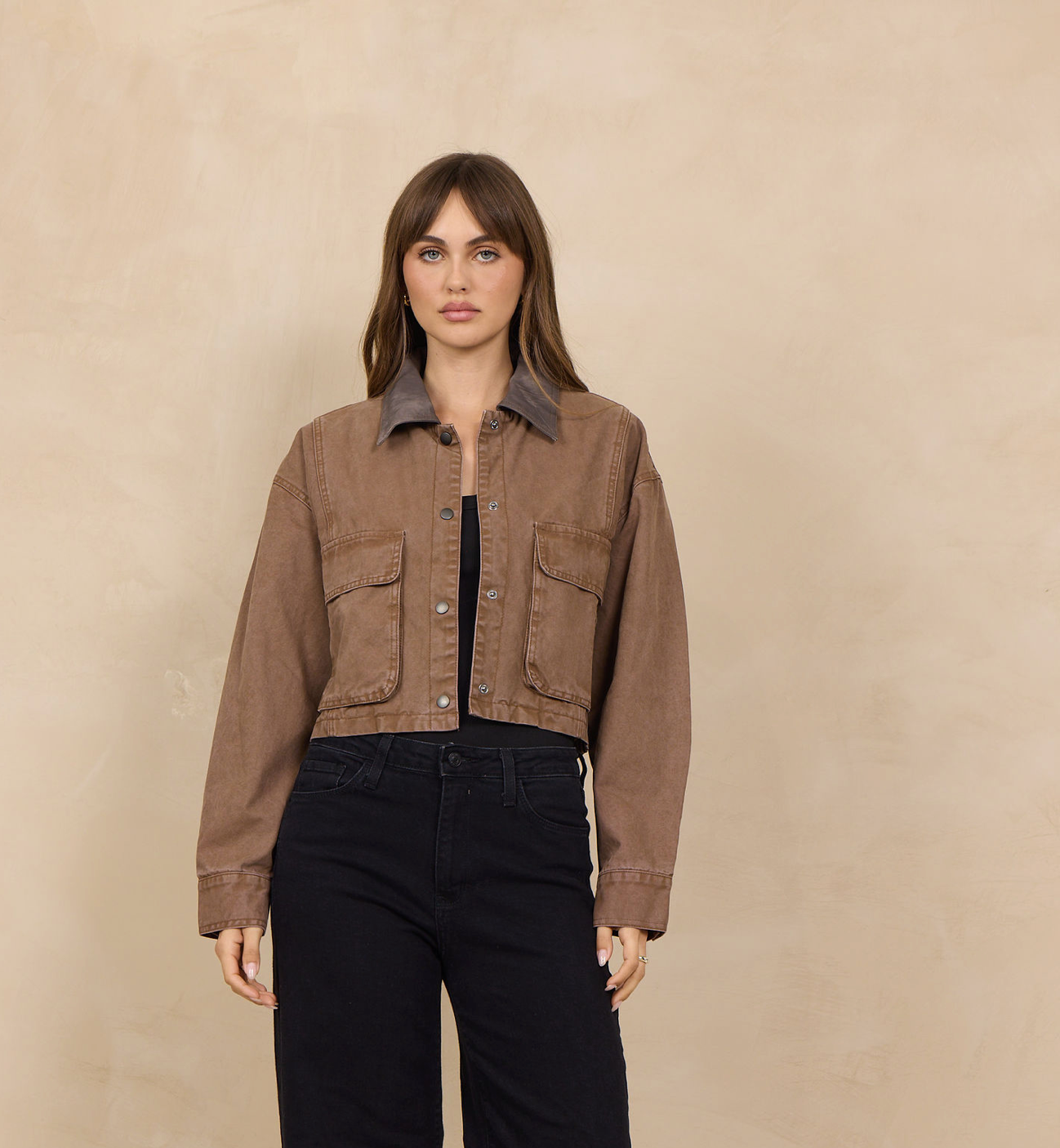 A woman stands against a warm beige textured wall wearing a cropped brown jacket with a contrasting dark collar, black wide-leg jeans, and black shoes.