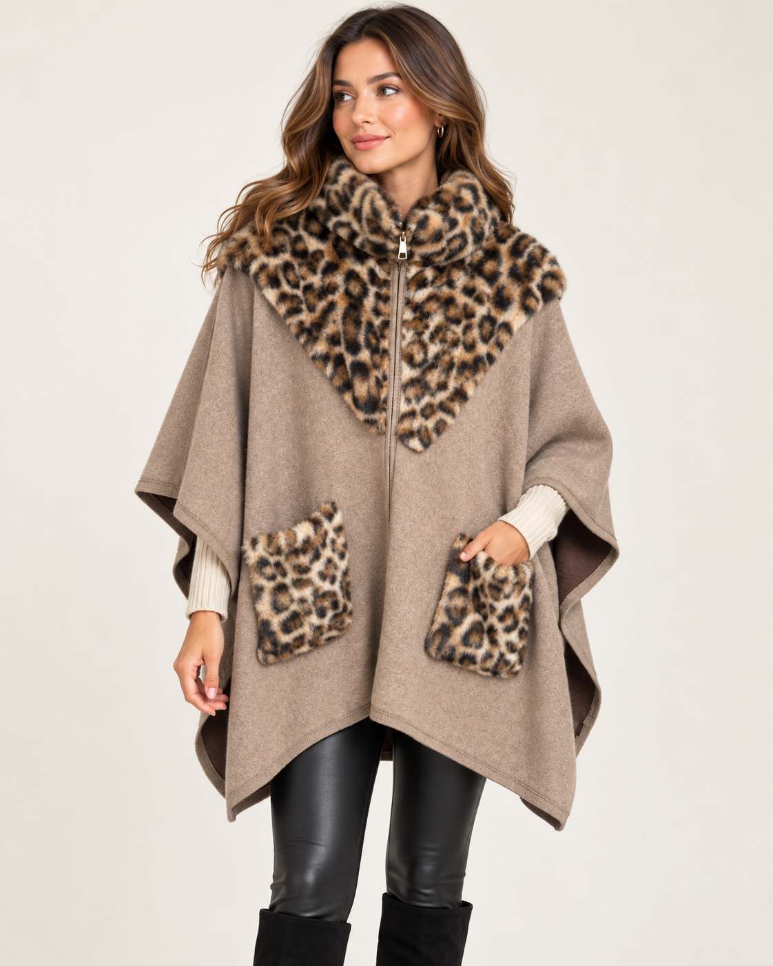 A woman models a taupe knit poncho with a large leopard-print faux fur collar and matching leopard-print pocket trims, paired with black leather-look pants and black boots against a light beige studio background.