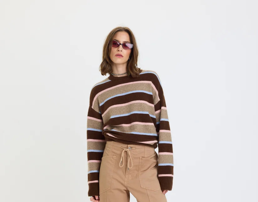 A model wearing a brown and beige striped knit sweater, tan drawstring trousers, and black loafers, standing against a plain white studio background.