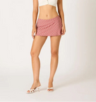A woman modeling a white short-sleeve crop top and a red-and-white gingham mini skirt, paired with white heeled sandals, against a plain light gray studio background.