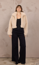 Model wearing a cropped faux fur jacket with an oversized collar, styled with a black tank and wide-leg black jeans against a neutral studio backdrop.