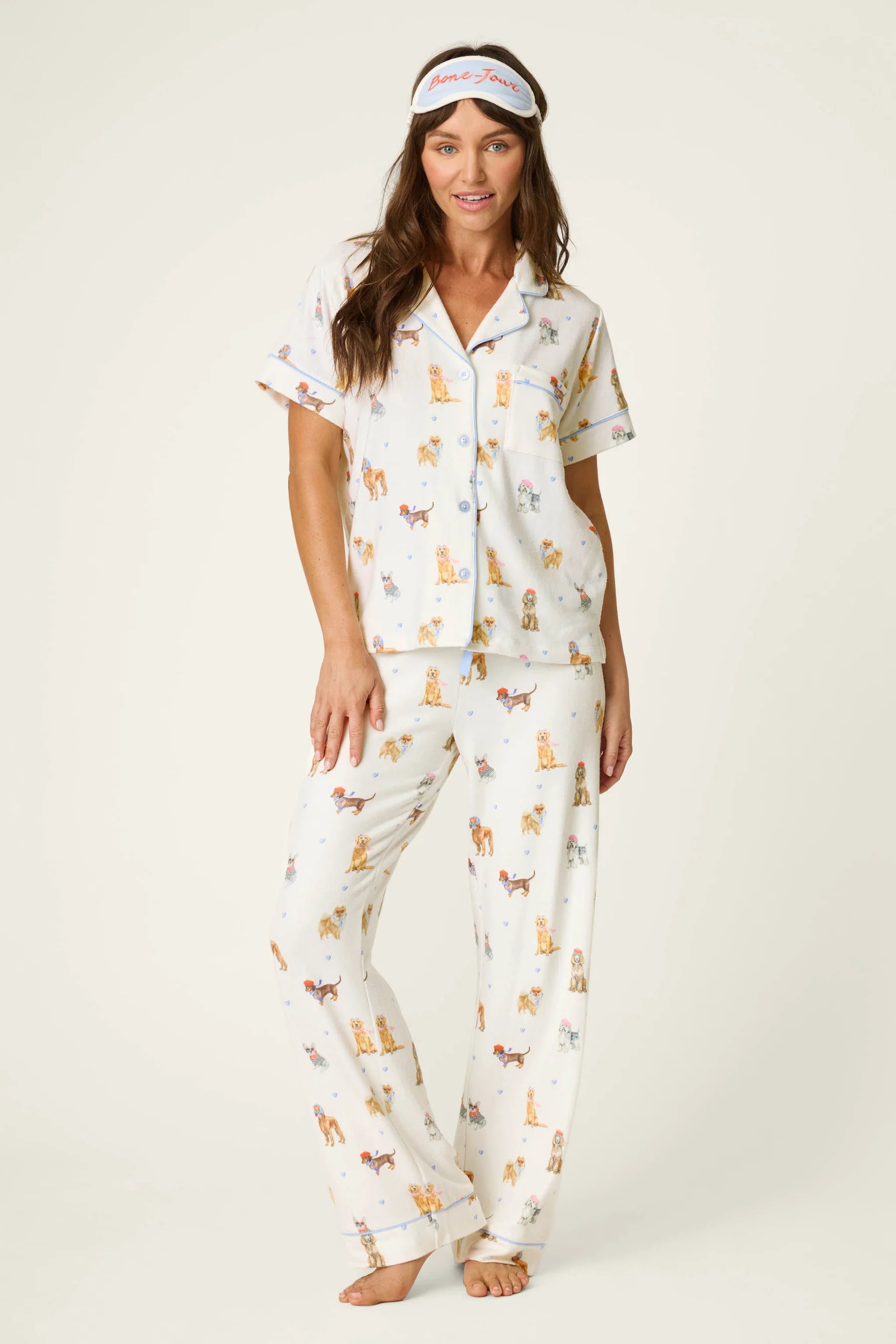 Woman wearing a three-piece dog-print pajama set with a button-front top, relaxed pants, and matching sleep mask.
