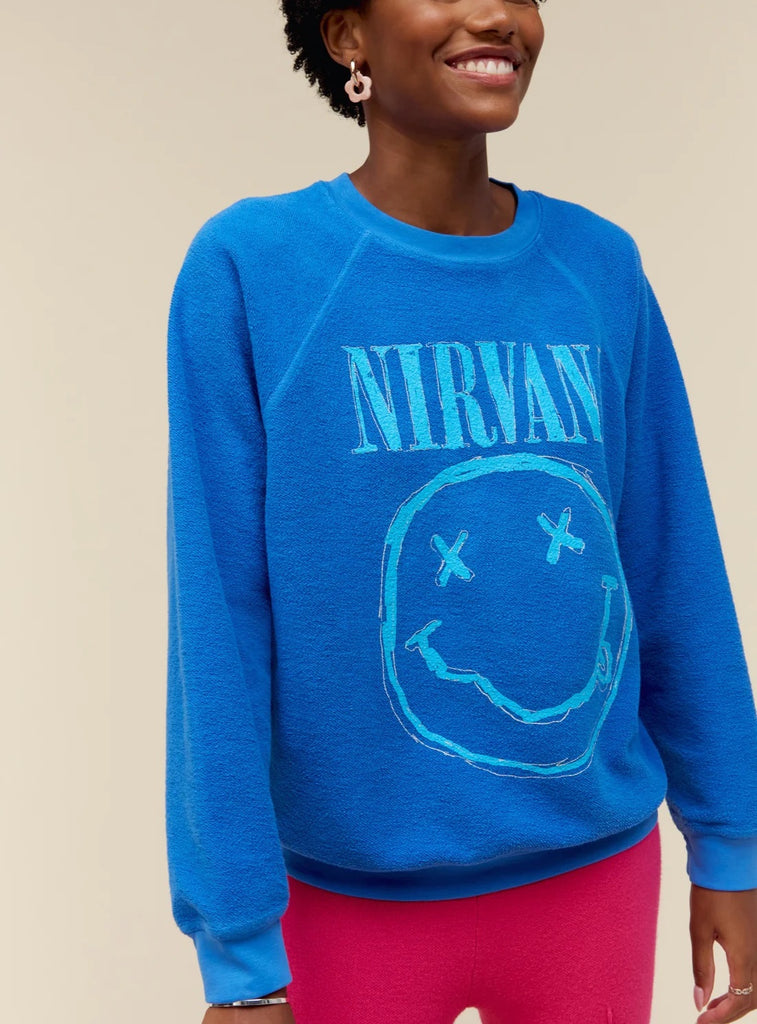 Day Dreamer Nirvana Smiley Crewneck Washed Cobalt. This crewneck features the smiley face logo on this reverse raglan crew in a sketch-like technique printed with a thicker ink to give it that hand painted look and feel. Designed to embody a vintage-find sweatshirt with a lightly brushed fleece and an intentional ‘inside out’ look for added novelty.