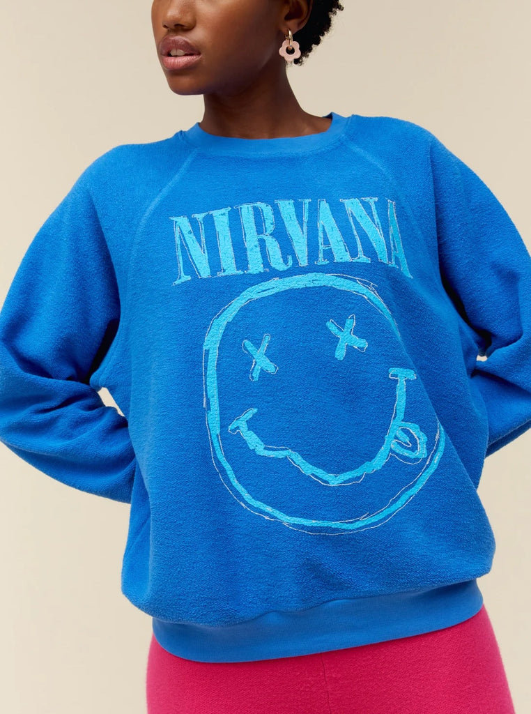 Day Dreamer Nirvana Smiley Crewneck Washed Cobalt. This crewneck features the smiley face logo on this reverse raglan crew in a sketch-like technique printed with a thicker ink to give it that hand painted look and feel. Designed to embody a vintage-find sweatshirt with a lightly brushed fleece and an intentional ‘inside out’ look for added novelty.
