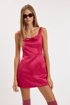 Model wearing a sleeveless mini dress with a square neckline, wide straps, a small bow at the neckline, and a gently flared silhouette.
