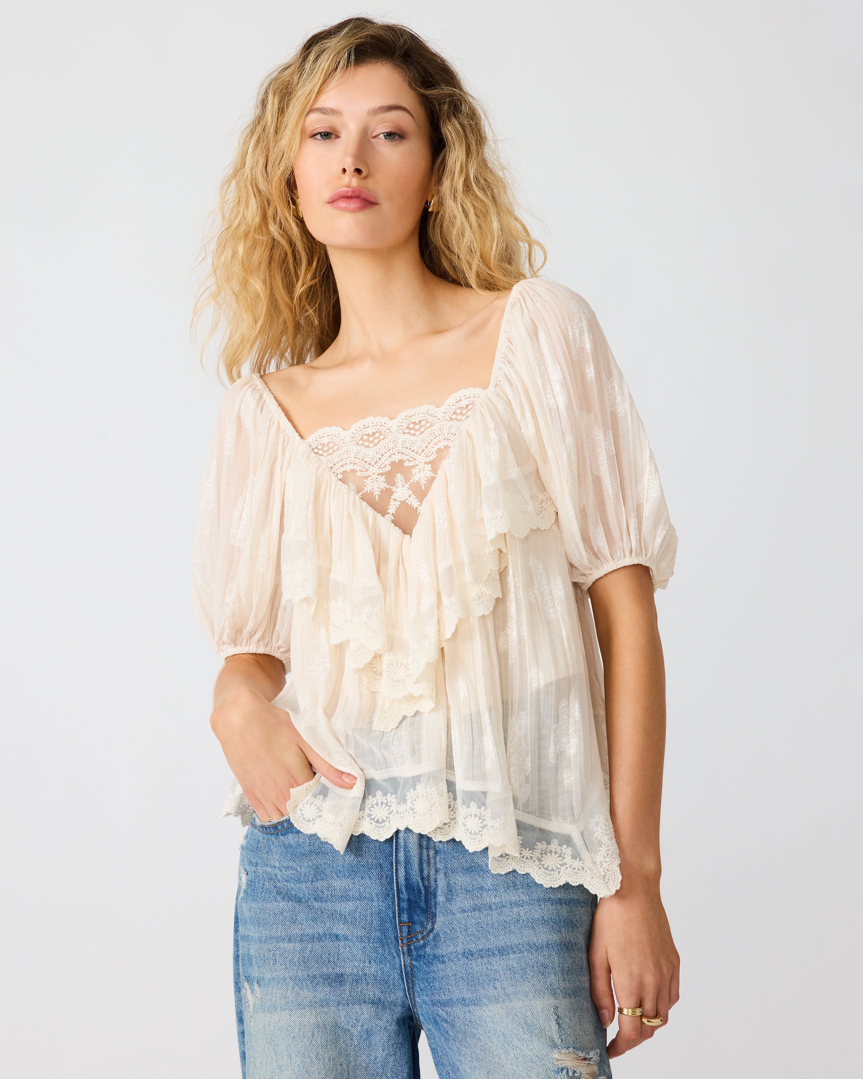 Model wearing a flowy cream lace blouse with ruffle details, puff sleeves, and a relaxed fit paired with blue denim jeans.