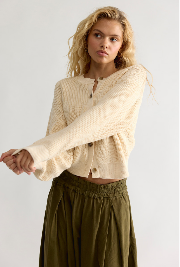 Model wearing a cropped button-front knit cardigan with a relaxed fit, ribbed trim, and long sleeves paired with a flowy skirt.