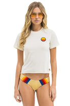 A woman modeling a white short-sleeve t-shirt with a small circular sunset graphic on the left chest, paired with yellow bikini bottoms featuring red, orange, and blue stripes. She is wearing aviator sunglasses with yellow lenses against a plain white background.