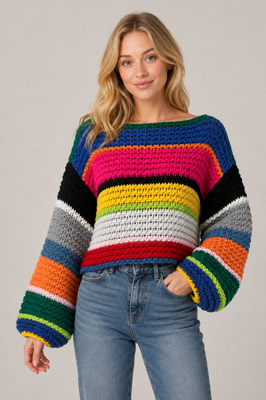 A woman wearing a chunky crocheted sweater with bold horizontal rainbow stripes in blue, pink, black, yellow, green, orange, white, and gray, paired with light-wash blue jeans, against a solid light gray background.