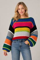A woman wearing a chunky crocheted sweater with bold horizontal rainbow stripes in blue, pink, black, yellow, green, orange, white, and gray, paired with light-wash blue jeans, against a solid light gray background.