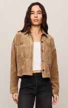A woman modeling a cropped tan suede jacket over a white tank top and black jeans, photographed against a plain light beige studio background.