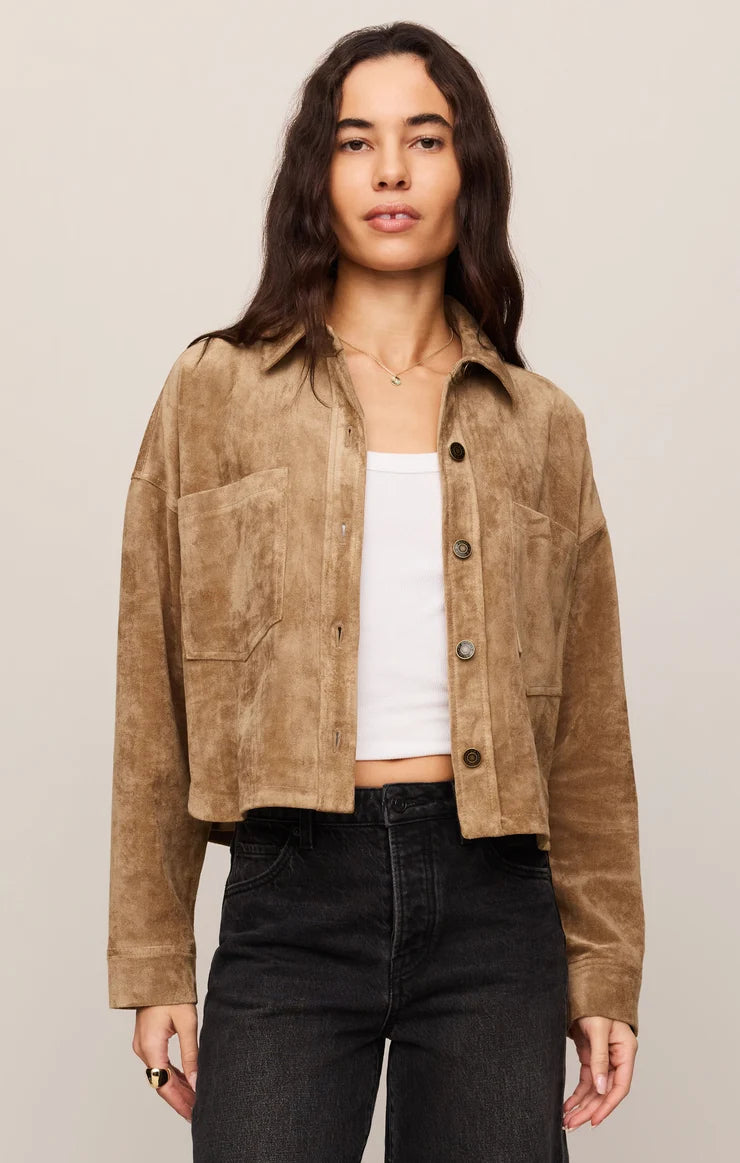 A woman modeling a cropped tan suede jacket over a white tank top and black jeans, photographed against a plain light beige studio background.