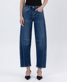 Model wearing high-rise barrel-leg jeans with seam detailing and a relaxed fit, styled with a black sleeveless top and pointed-toe heels against a white studio background.