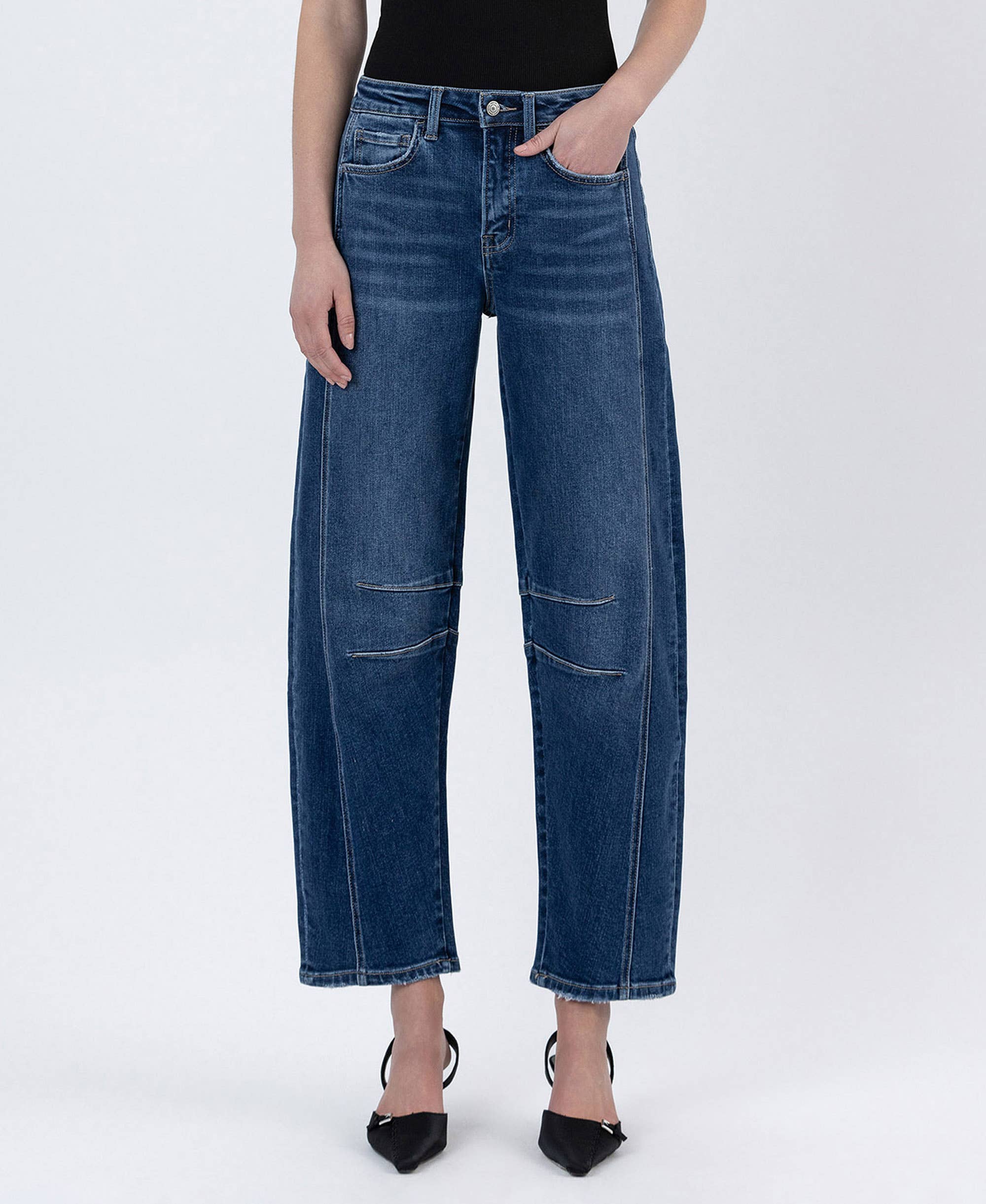 Model wearing high-rise barrel-leg jeans with seam detailing and a relaxed fit, styled with a black sleeveless top and pointed-toe heels against a white studio background.