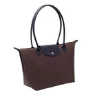 A brown and black leather tote bag with double shoulder straps, displayed on a white table next to a stack of books in front of a white curtain background.