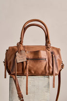 Leather satchel handbag with dual top handles, multiple exterior zip pockets, and a removable crossbody strap displayed on a pedestal against a neutral background.