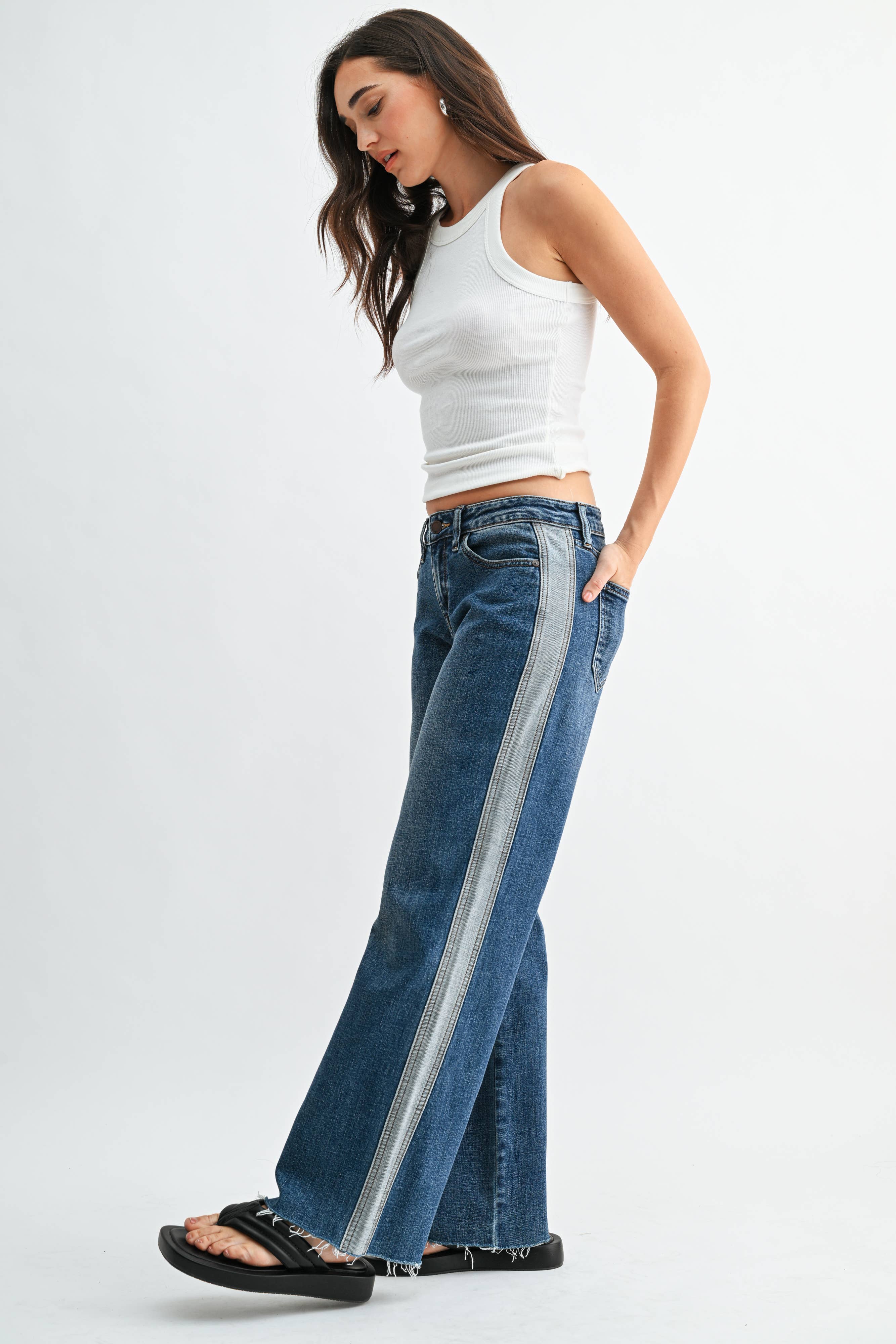 Model wearing high-rise wide-leg jeans with contrast side panel detailing, five-pocket styling, and a raw hem paired with a fitted ribbed tank.