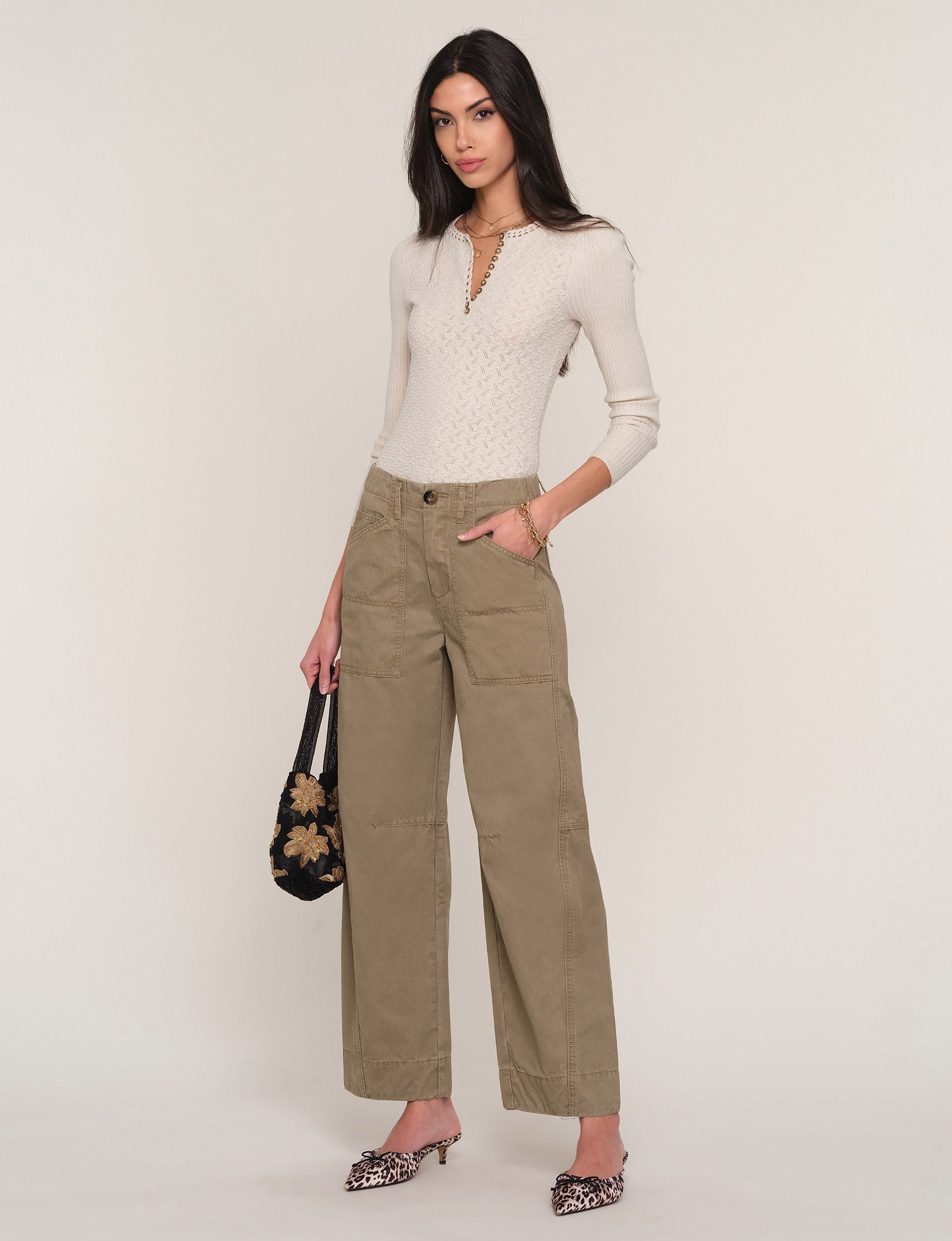 A model stands against a light beige studio background wearing a cream-colored textured knit top, olive green wide-leg cropped pants, and leopard-print pointed-toe mules. She is holding a black floral-patterned handbag.