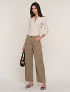 A model stands against a light beige studio background wearing a cream-colored textured knit top, olive green wide-leg cropped pants, and leopard-print pointed-toe mules. She is holding a black floral-patterned handbag.
