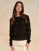 A woman modeling a black textured knit sweater with a crew neck and long sleeves, paired with cream-colored pants, set against a warm beige textured background.