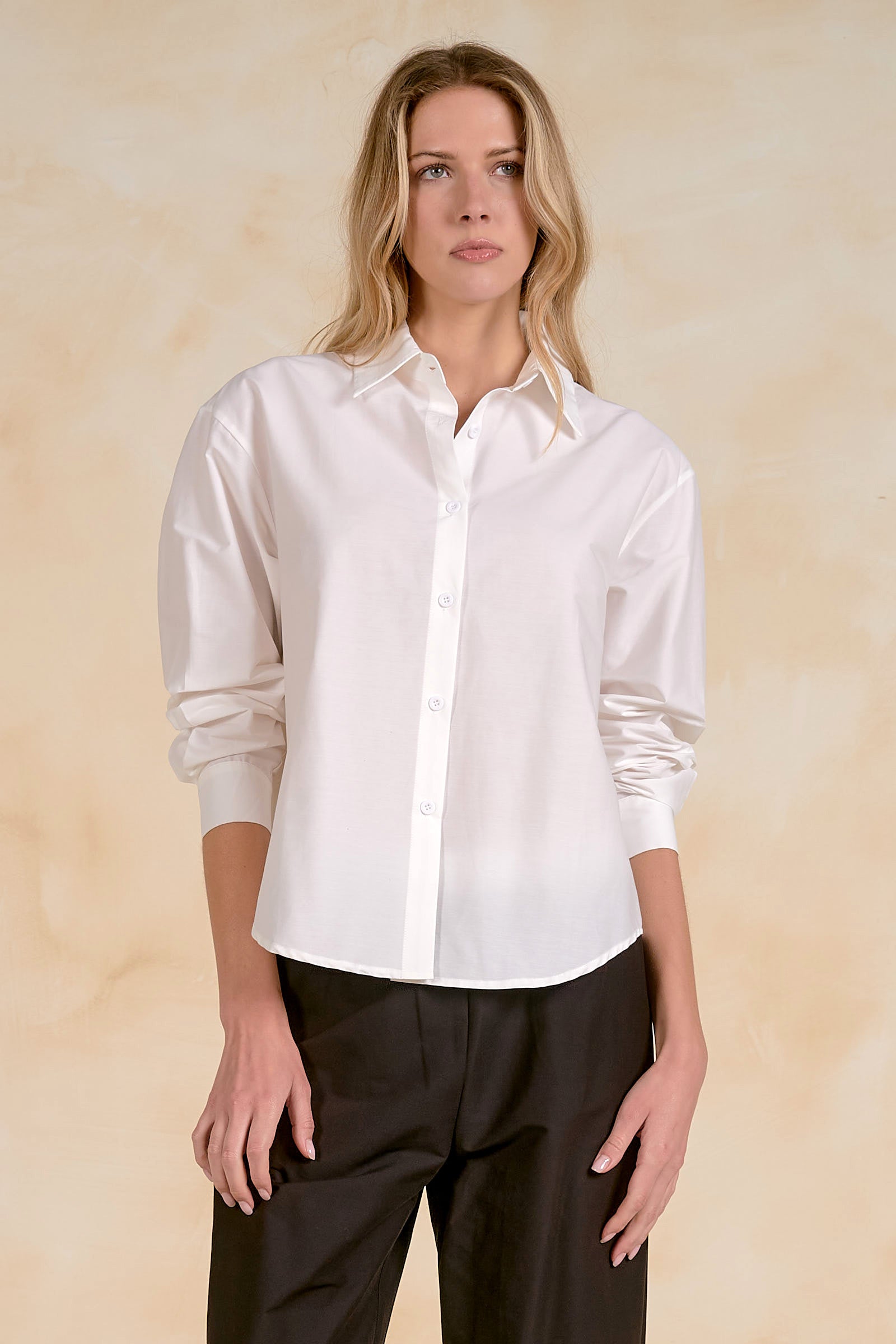 Model wearing a classic white button-front shirt with a collared neckline, long sleeves, and a relaxed fit paired with tailored trousers.
