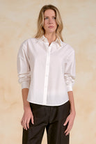 Model wearing a classic white button-front shirt with a collared neckline, long sleeves, and a relaxed fit paired with tailored trousers.