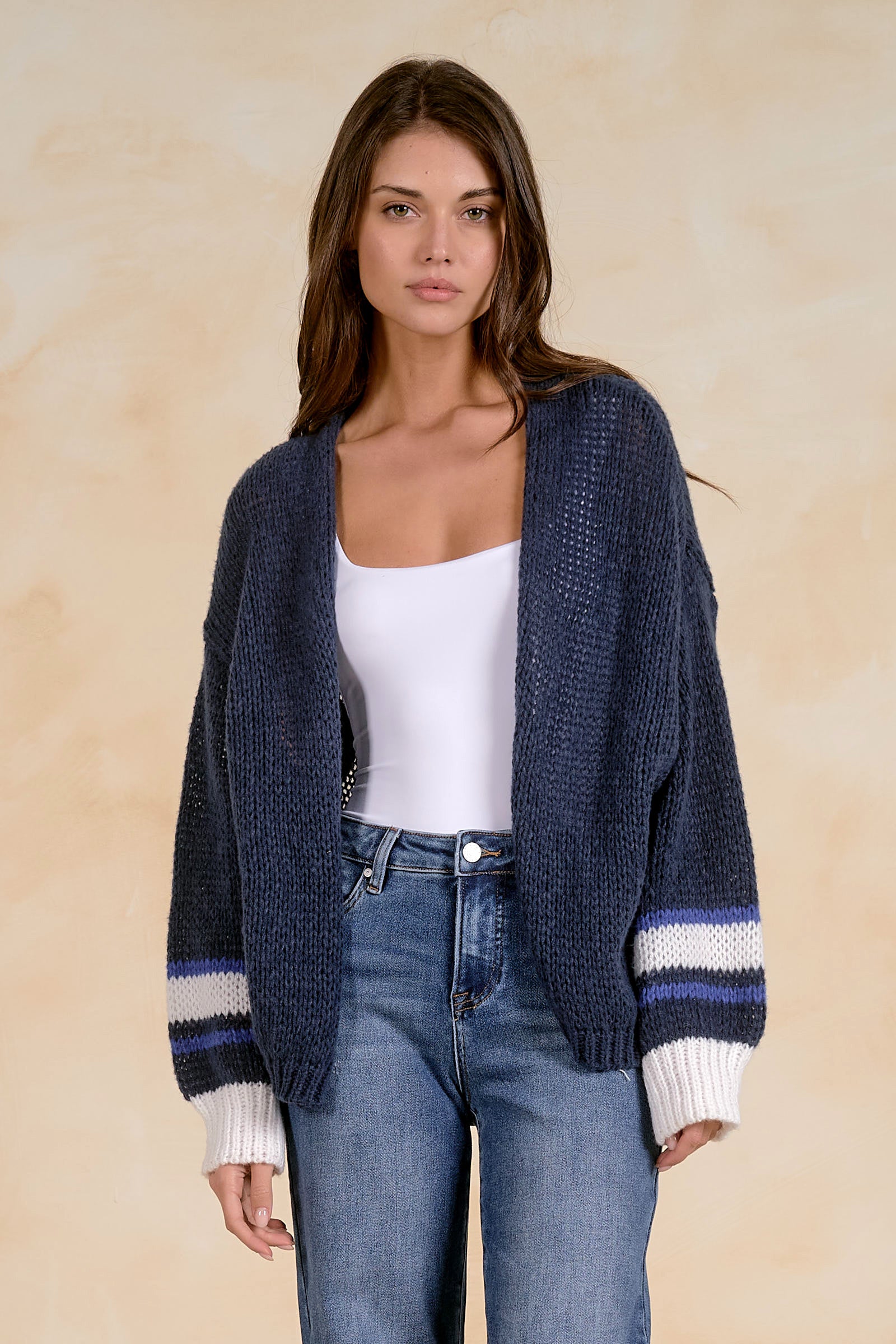 Model wearing an oversized chunky knit open-front cardigan with striped ribbed cuffs layered over a fitted tank and blue denim jeans.