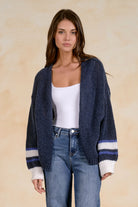 Model wearing an oversized chunky knit open-front cardigan with striped ribbed cuffs layered over a fitted tank and blue denim jeans.