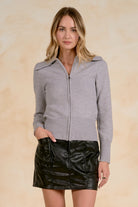 Model wearing a fitted zip-front knit cardigan with a wide collar, ribbed trim, and long sleeves paired with a faux leather mini skirt.