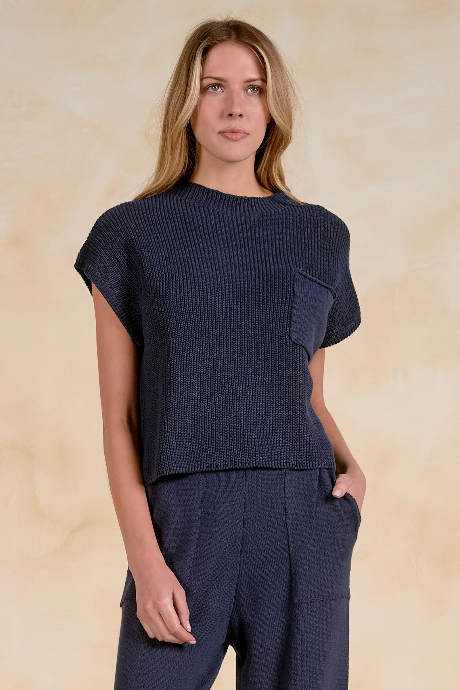 Model wearing a textured cap sleeve knit sweater with a crew neckline, relaxed fit, front patch pocket, and ribbed trim, paired with coordinating knit pants.