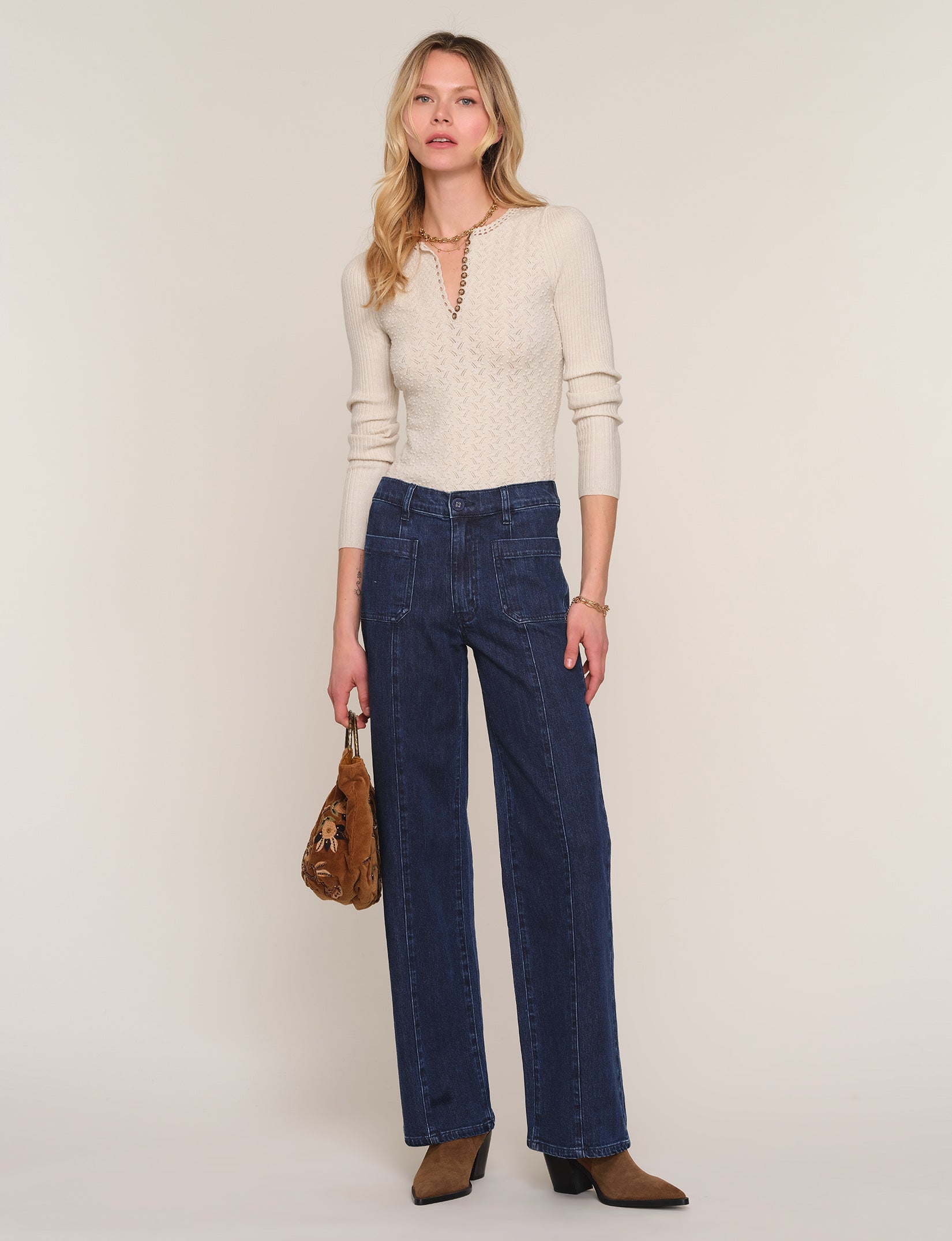 A woman modeling a cream-colored knit long-sleeve top with a keyhole cutout and dark blue wide-leg jeans, holding a brown textured handbag against a solid light beige studio background.